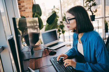 Businesswoman at desk working with computer monitor and laptop in modern loft office focused on coding projects