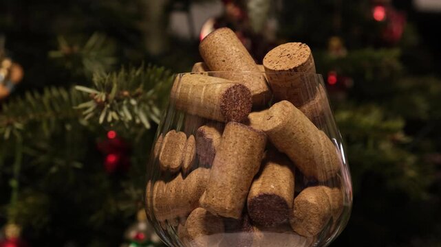 An overflowing glass with red wine corks and champagne stopper on the background of a Christmas tree. Concept of alcohol abuse during the Christmas holidays. 4K video, turntable, glass rotating.