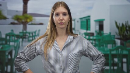 Woman in striped blouse gesturing hand on outdoor restaurant terrace with green chairs and white building facade; confidence.