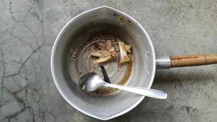 Photo from above of a dirty pot containing bones, spoon and food scraps lying on a cement floor