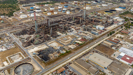 Fototapeta premium Aerial view of a large, complex oil and chemical refinery. The facility features a tall chimney, dense industrial piping and machinery, and a vast storage area of cylindrical tanks.
