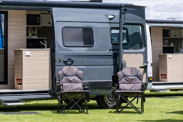 Modern camper van with open sliding doors and two folding camping chairs set up on green grass in an outdoor camping area. Concept of van life, outdoor leisure, road trip lifestyle, mobile living