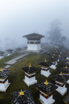 Aerial view of white chortens with dark roofs rising through the mist, a serene landscape at Druk Wangyal Chortens, Dochula Pass, Bhutan.