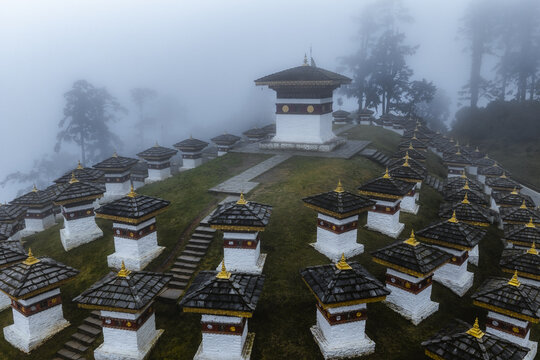 Aerial view of the Druk Wangyal Chortens shrouded in mist, their white walls and golden roofs contrasting with the green landscape, Dochula Pass, Bhutan.