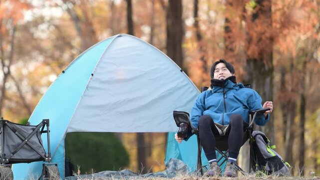 秋のキャンプで椅子に座りリラックスする男性ソロキャンパー、アウトドア楽しい～景色を眺めるリラックスして伸びをするクローズアップの動画　落ち葉の前ボケあり
