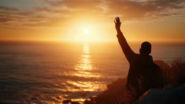 Silhouette of a person on a cliff raising an arm toward a golden sunset over the ocean.