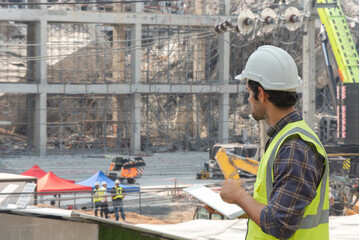 Young male engineer wearing safety vest and hard hat using a digital tablet to inspect the construction site. Building under construction and crane in background.