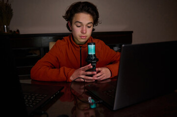 A young person in an orange hoodie sits at a desk with a glowing blue microphone and laptop, creating content in a dim roo
