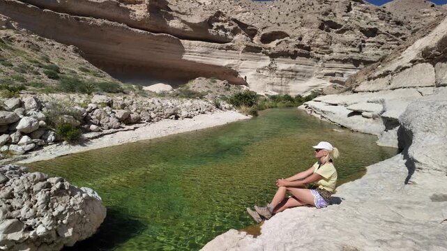 Woman wearing a cap sitting by the clear green water of a natural oasis, surrounded by rugged rock formations and desert vegetation in tranquil wadi environment at Wadi Hadhbram in Oman in Dhofar