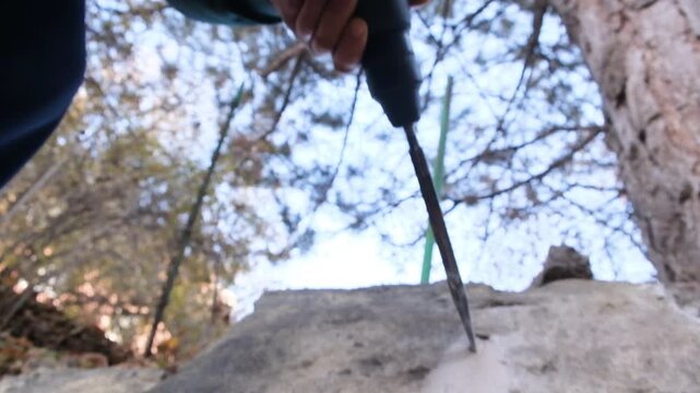 Low-angle wide shot of a worker using a powerful hammer drill with a flat chisel bit to break concrete. Dust from the fractured concrete falls down the stone surface.