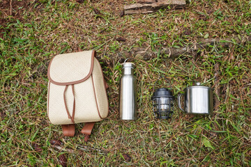 A set of outdoor gear including a woven rattan backpack, a electric lantern, a stainless steel water bottle, and a stainless steel mug, placed on a grassy surface with a background of pine tree trunks