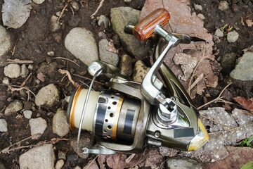A metal fishing reel with a wooden handle placed on the ground by the riverbank.