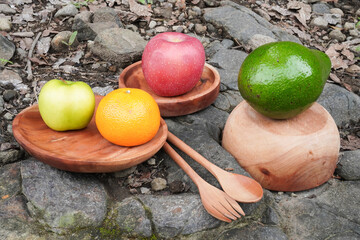 Fruits such as avocado, apple, and orange placed on wooden plates and bowls with a rocky background during outdoor activities.