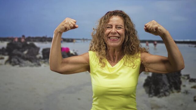 Middle age hispanic woman flexing both biceps at beach in yellow sleeveless top, smiling with raised arms; confidence healthy aging.
