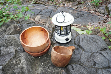 A white portable hanging electric camping lantern with wooden plates, bowls, and mugs placed on soil and rocky ground during outdoor activities.