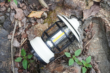 A white portable hanging electric camping lantern placed on soil and rocky ground during outdoor activities.