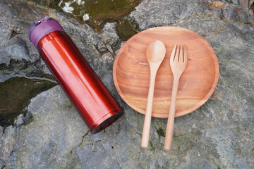 A red insulated tumbler with a wooden plate, spoon, and fork placed on a stone surface.