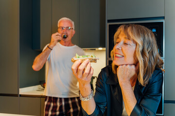 Senior couple starts their day with a healthy breakfast in a bright kitchen, the woman smiling as she holds toast with avocado and kiwi while the man drinks coffee