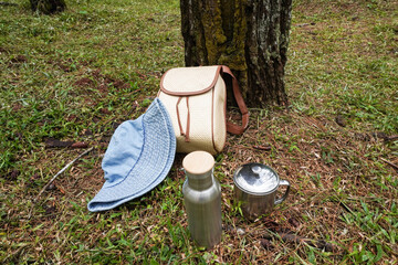A set of outdoor gear including a woven rattan backpack, a bucket hat, a stainless steel water bottle, and a stainless steel mug, placed on a grassy surface with a background of pine tree trunks.
