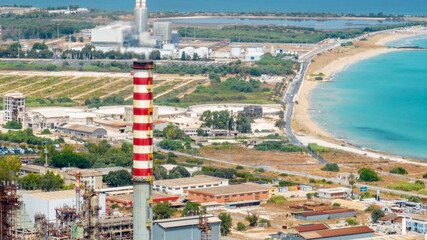A coastal industrial complex dominated by a tall, red and white striped smokestack emitting smoke. The refinery contrasts with the background featuring a road, green fields, and a bright turquoise sea