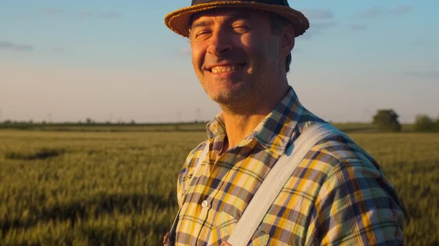 Portrait of smiling satisfied farmer whit hat standing in wheat field at sunset looking at camera.