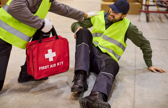 Warehouse worker injured in accident gets firstaid. A helper kneels with a first aid kit, giving emergency care for injury and rescue on the warehouse floor. Clear safety and rescue concept.