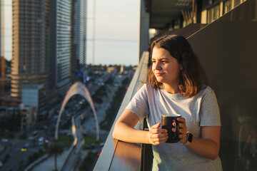 Woman enjoying a warm drink on a balcony during golden hour at sunrise or sunset, looking at the...