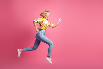 Young woman in yellow shirt and jeans leaps against pink background showcasing casual fashion energy
