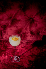 A pale cocktail with an orange slice garnish in a coupe glass, set against a luxurious, deep red tufted velvet background. Mood lighting creates a dramatic shadow