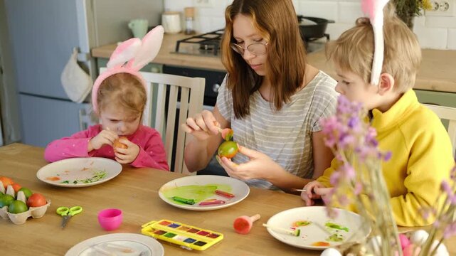 Children siblings painting Easter egg sitting together at table. Easter Family traditions. Teen girl teaching happy little brother and sister to dye, decorate eggs with paints. Preparation to holidays