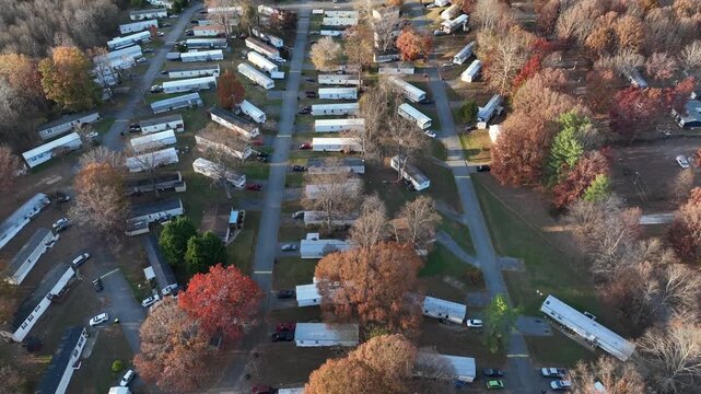 Aeria flyover large American mobile trailer home park set among autumn trees as long rows of white units line narrow roads and quiet community. Wooded landscape in soft daylight in american. top down