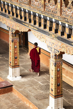 Aerial view of a solitary monk in vibrant maroon robes walking under ornately carved wooden beams, a symphony of golden hues and intricate details, Punakha, Bhutan.