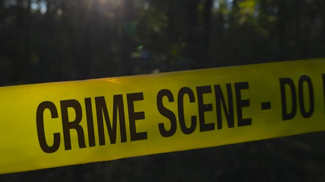 Cinematic close up video moving around and in front of a yellow forensic sciene police crime scene tape ribbon that is pulled in front of a forest area while sunlight backlights the fluorecent tape.