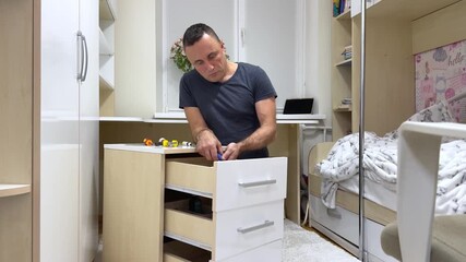 man works on fixing a white cabinet in a childrens room focusing on its details and making careful adjustments creating a calm practical home improvement moment filled with gentle family atmosphere.