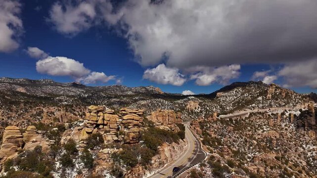 Aerial view of the winding Mount lemmon road cuts through the rugged landscape with snow-dusted mountains under a sky of bright blue and billowing clouds, Tucson, Arizona, United States.