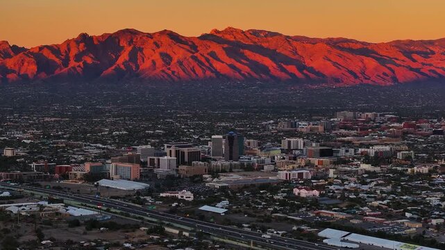 Aerial view of Tucson showing the city buildings and mountains under a vibrant sunset, with warm orange hues contrasting cool shadows, Tucson, Arizona, United States.