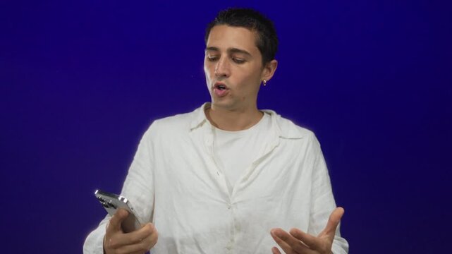 Hispanic man in white shirt interacting with smartphone over isolated blue background, expressing emotions and gestures, capturing technology use in modern communication.