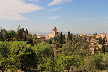 A panoramic view of the Alhambra surrounded by lush greenery, with historic stone buildings and a church tower rising above the trees.