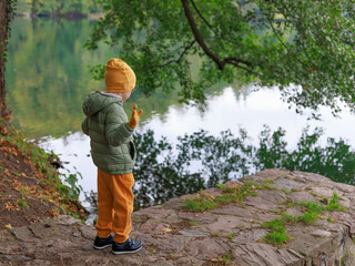 A child in autumn clothes stands on the lakeside path, turning away and raising a hand as if...