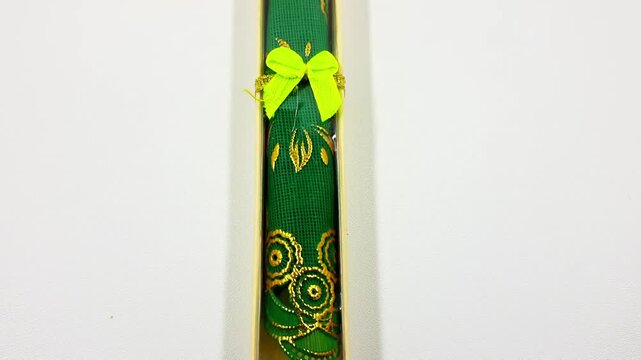 Green fabric scroll featuring intricate gold embroidery and a small neon yellow bow sits vertically packaged against a stark white background suggesting a cultural gift or decorative keepsake item.