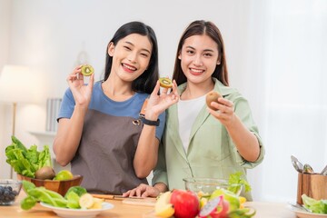 Two women hold kiwis and smile while preparing food in a bright kitchen during daytime, with various fruits and vegetables on the counter
