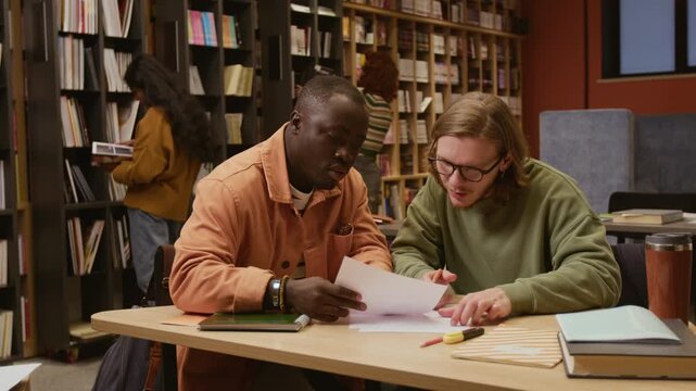 Young adult African American male student discussing test sheets with groupmate while collaborating on academic task at library table