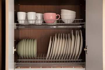 Open kitchen cabinet with neatly arranged plates, bowls and cups on metal drying racks. Organized dish storage, clean minimalist home interior with pastel tableware.