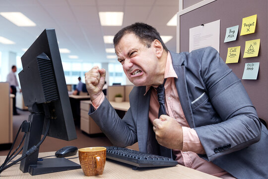 A stressed businessman in a gray suit clenches his fists at a computer screen in a busy office, showing frustration.