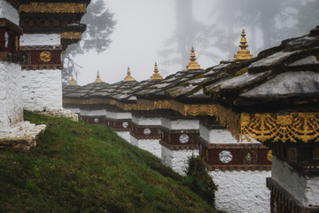 View of white chortens with golden roofs stand in misty Dochula Pass, their ornate details a stark contrast to the soft green hillside, Hungtso, Bhutan.