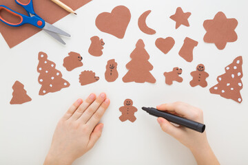 Little child hand holding marker and drawing smiling face on brown gingerbread man shape. Light gray table background. Making Christmas decoration from application paper. Closeup. Top down view.