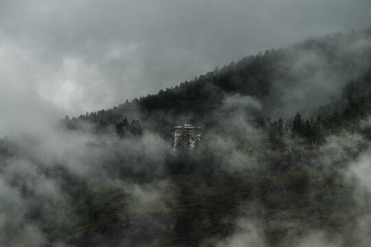 View of a building shrouded in mist, amidst a dense forest clinging to the steep slopes under a brooding sky, evoking mystery and serene isolation, Paro, Bhutan.