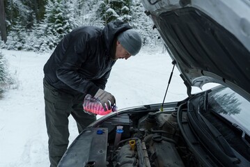 A man is standing under the open hood of a car and pouring antifreeze . Winter car care. Car maintenance in winter.