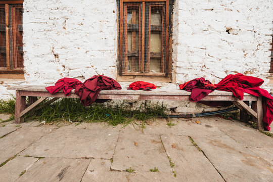 View of vibrant red robes resting on a weathered wooden bench against a rustic white stone wall with aged wooden window frames, Paro, Bhutan.