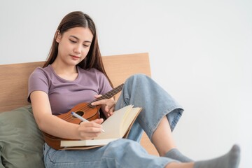Young woman plays ukulele and writes in notebook while sitting on bed in cozy room during daytime